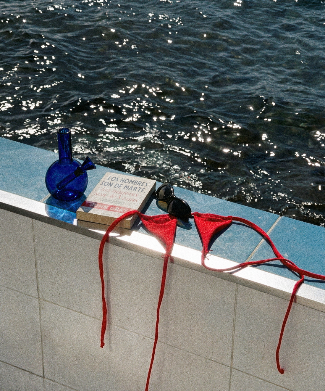 loke glass gaia in bvlue next to Red bikini straps, blue bottle, and book on a ledge with water in the background